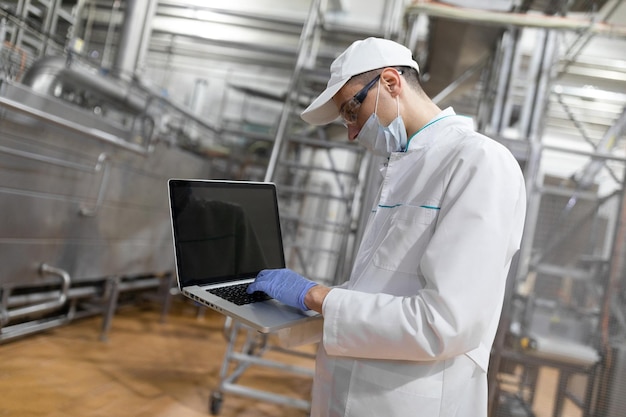 Technologist in a white coat mask and gloves with a laptop in his hands is in the shop for the production of butter and cheese production process at the dairy plant