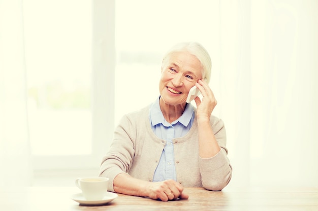 technology, communication age and people concept - happy senior woman with smartphone and coffee sitting at table and calling at home