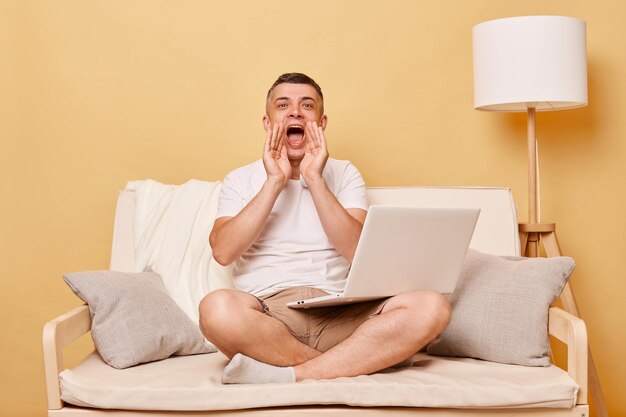 Technology communication Computer technology Online user Positive young man working on laptop computer sitting on comfortable couch at home screaming making announcement