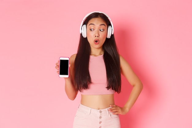 Technology, communication and online lifestyle concept. Excited and happy asian girl looking amazed, showing smartphone screen while listening podcast or music in headphones, pink wall.