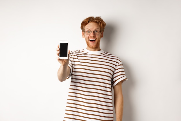 Technology and e-commerce concept. Happy young redhead man in glasses showing blank smartphone screen, looking at camera amazed, standing over white background