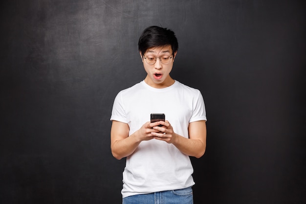 Technology, gadgets and people concept. Amazed and impressed handsome asian guy react to exciting news he read online, holding mobile phone, stare smartphone screen amused