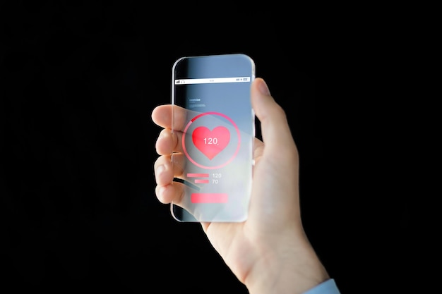 technology, health care, application and people concept - close up of male hand holding and showing transparent smartphone with red heart icon on screen and measuring pulse over black background