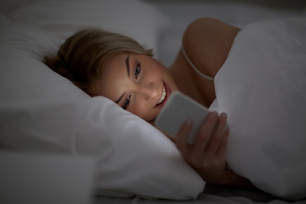 technology, internet, communication and people concept - happy smiling young woman texting on smartphone in bed at home bedroom at night