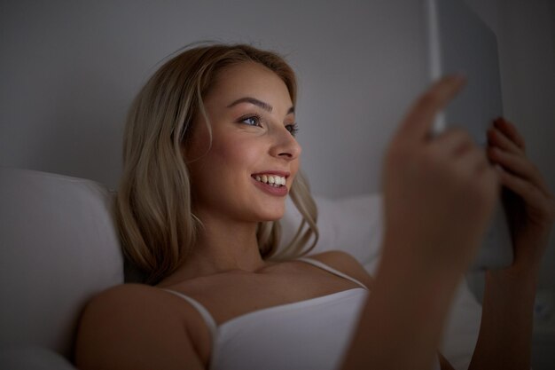 technology, internet, communication and people concept - happy smiling young woman with tablet pc computer in bed at home bedroom at night