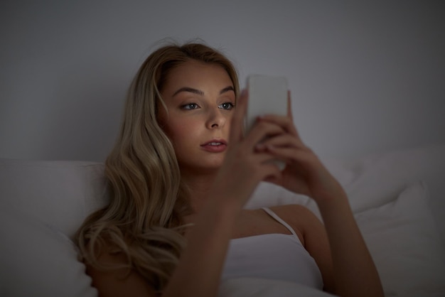 technology, internet, communication and people concept - young woman texting on smartphone in bed at home bedroom at night