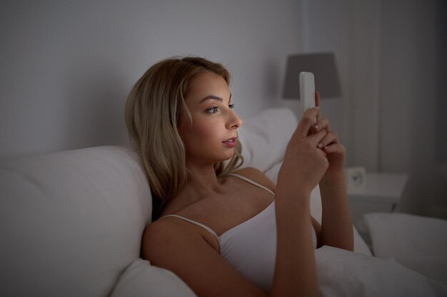 technology, internet, communication and people concept - young woman texting on smartphone in bed at home bedroom at night