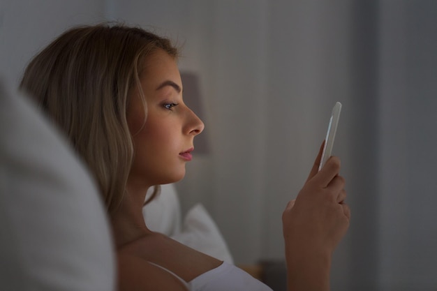 technology, internet, communication and people concept - young woman texting on smartphone in bed at home bedroom at night