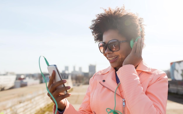 technology, lifestyle and people concept - smiling african american young woman or teenage girl with smartphone and headphones listening to music outdoors