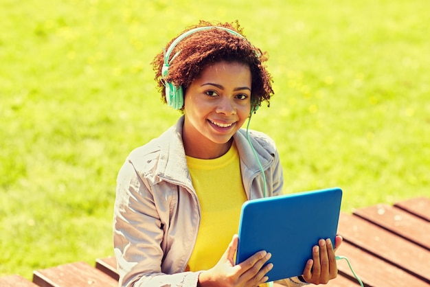 technology, lifestyle and people concept - smiling african american young woman or teenage girl with tablet pc computer and headphones listening to music in summer park