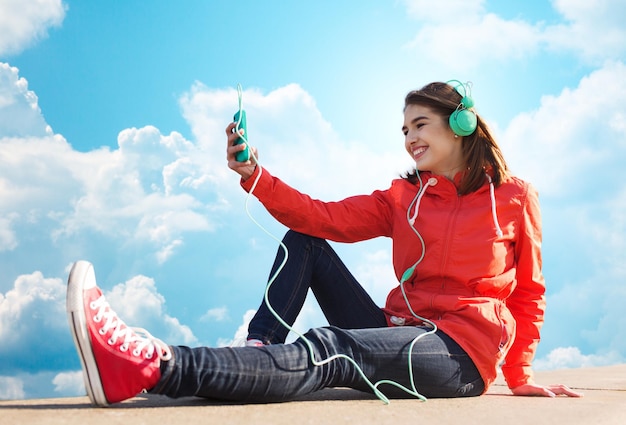 technology, lifestyle and people concept - smiling young woman or teenage girl with smartphone and headphones listening to music outdoors over blue sky and clouds background