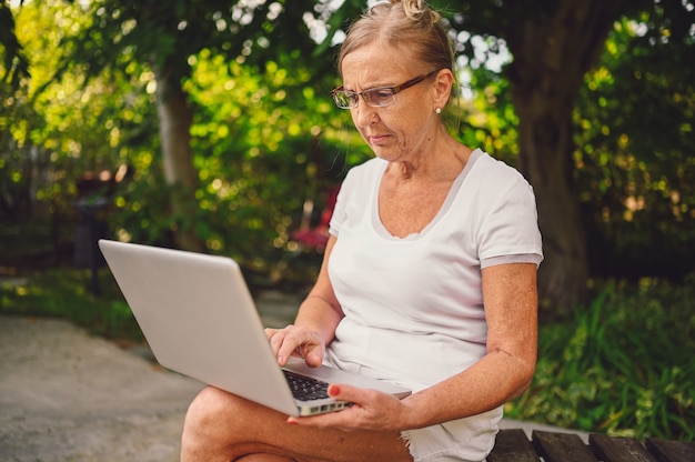 Technology, old age people concept - elderly happy senior old woman working online with laptop computer outdoor in the garden. Remote work, distance education.