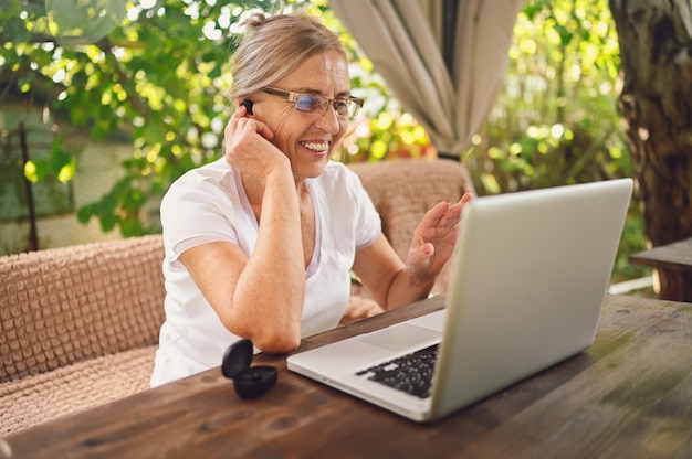 Technology, old age people concept - elderly happy senior woman using wireless headphones working online with laptop computer outdoor in the garden. Remote work, distance education.