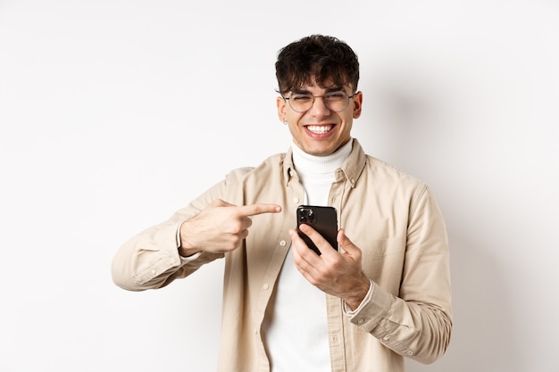 Technology and online shopping concept. Portrait of natural man in glasses laughing and pointing finger at smartphone, showing promo offer on screen, standing white background