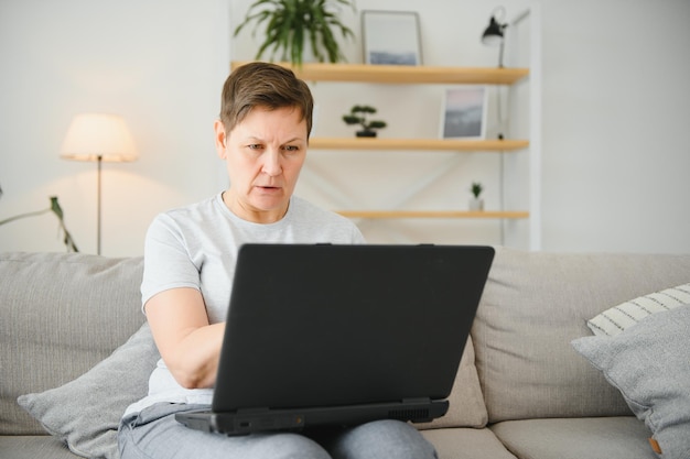 Technology people and communication concept Happy senior woman with tablet pc computer having video chat at home Modern middleaged 50s grandmother sit relax on couch in living room using laptop