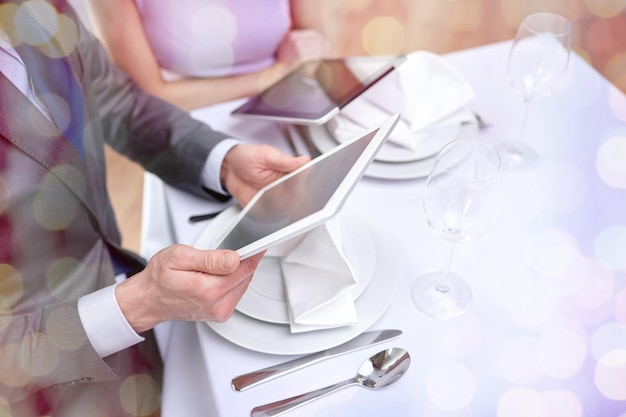 technology, people, food and holiday concept - close up of couple with menus on tablet pc computers at restaurant over holidays lights background