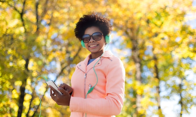 technology, season and people concept - smiling african american young woman or teenage girl with smartphone and headphones listening to music over autumn park background