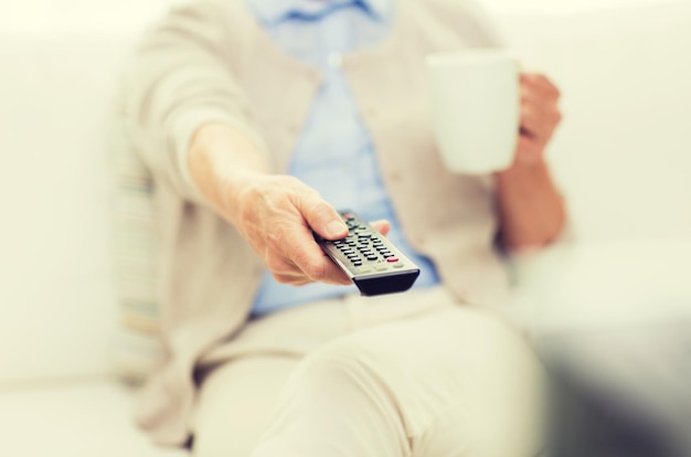 technology, television, age and people concept - close up of senior woman watching tv, drinking tea and changing channels by remote control at home