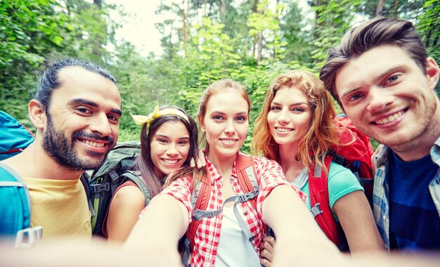 technology, travel, tourism, hike and people concept - group of smiling friends walking with backpacks taking selfie by smartphone or camera in woods