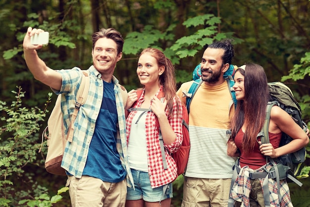 technology, travel, tourism, hike and people concept - group of smiling friends walking with backpacks taking selfie by smartphone in woods