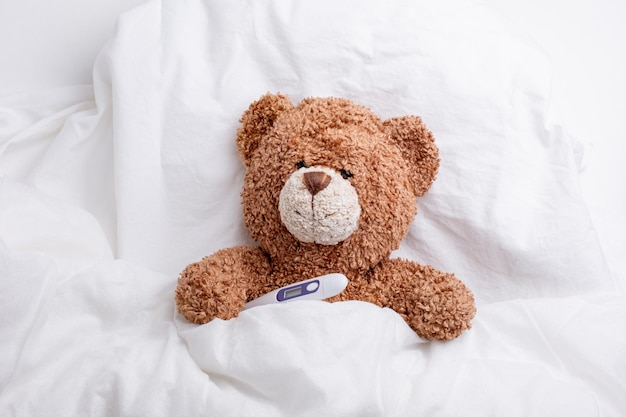 Teddy bear is lying in bed under a blanket with a thermometer white background the concept medicine