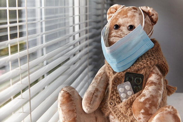 A teddy bear in a medical protective mask and with pills in his pocket looks wistfully through a rainy window