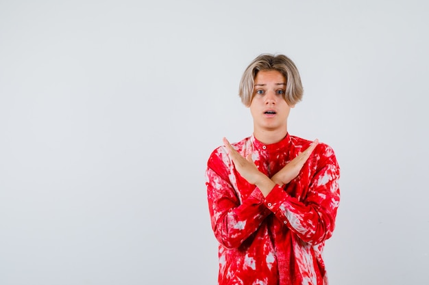 Teen blonde male in oversize shirt showing stop gesture with crossed hands and looking bewildered , front view.