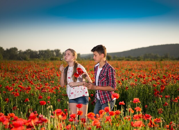 Teen boy and girl posing on the poppy field at sunset.Gilr blowing blowball. Summer sunny day