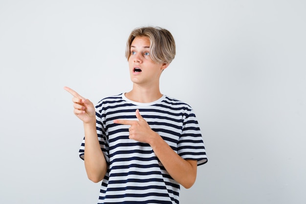 Teen boy pointing to the left side, opening mouth in t-shirt and looking shocked , front view.