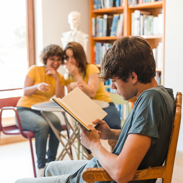 Teen boy reading book near gossiping classmates