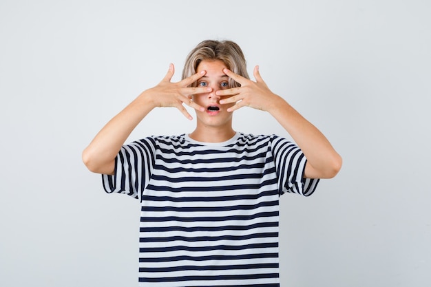 Teen boy in t-shirt looking through fingers and looking surprised , front view.