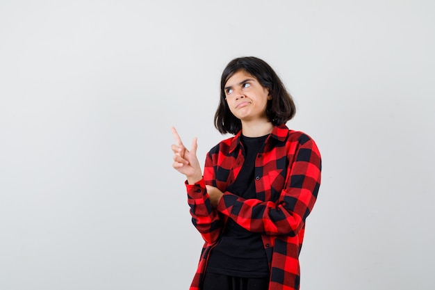 Teen girl in casual shirt pointing up, looking away and looking pensive , front view.