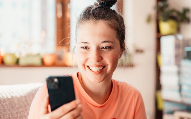 Teen girl checking social media holding smartphone at home Smiling young woman using mobile phone app playing game shopping online