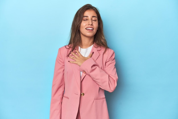 Teen girl in chic pink suit on a blue background laughs out loudly keeping hand on chest