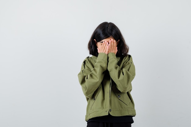 Teen girl covering face with hands in t-shirt, green jacket and looking sorrowful. front view.