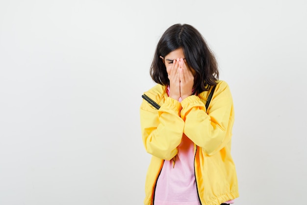 Teen girl covering face with hands in t-shirt, yellow jacket and looking focused , front view.