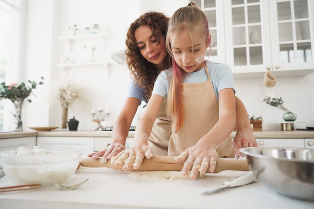 Teen girl helping her mom to cook dough in their kitchen at home