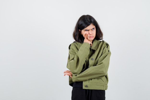 Teen girl holding finger on cheek, looking away, frowning face in army green jacket and looking upset. front view.