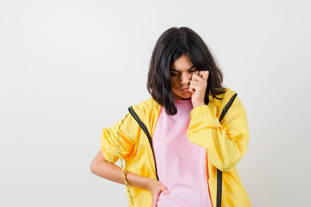 Teen girl holding hand on cheek and waist in yellow tracksuit, t-shirt and looking thoughtful , front view.