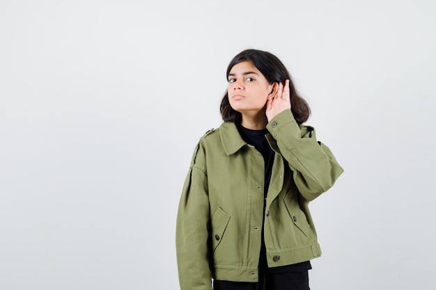 Teen girl holding hand behind ear in green jacket and looking serious , front view.