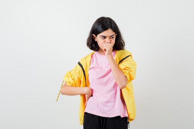 Teen girl holding hand on mouth and waist in yellow tracksuit, t-shirt and looking puzzled , front view.