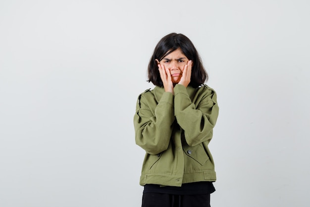 Teen girl holding hands on cheeks in t-shirt, jacket and looking sorrowful , front view.