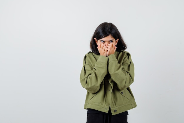 Teen girl holding hands on mouth in t-shirt, jacket and looking cowardly. front view.