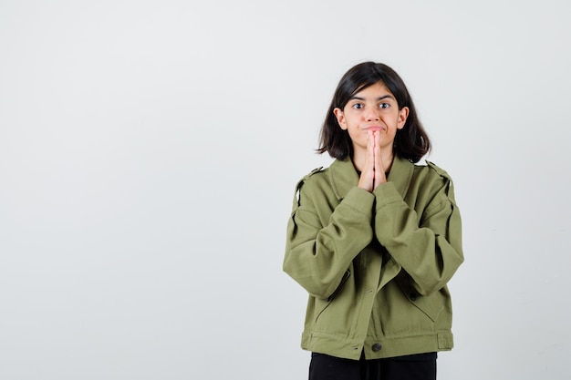 Teen girl holding hands in praying gesture in army green jacket and looking focused , front view.