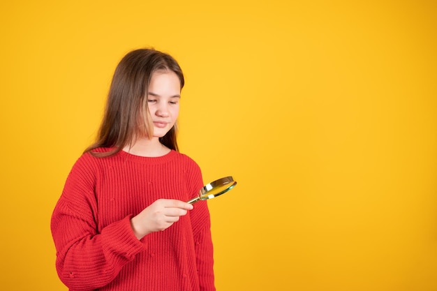 Teen girl holding magnifying glass and looking at something Young schoolgirl in red sweater on yellow background