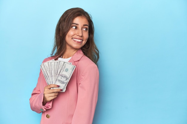 Teen girl in pink flaunting a handful of dollar bills looks aside smiling cheerful and pleasant