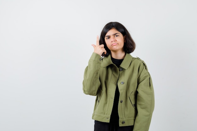 Teen girl pointing up in army green jacket and looking dismal , front view.