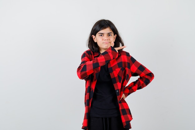 Teen girl showing gun gesture in casual shirt and looking puzzled , front view.