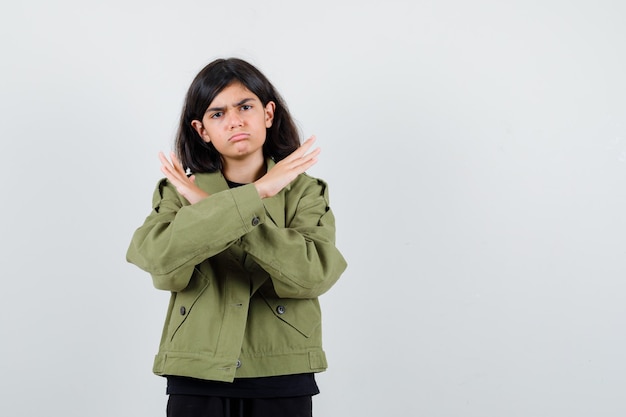 Teen girl showing refusal gesture in t-shirt, green jacket and looking disappointed , front view.