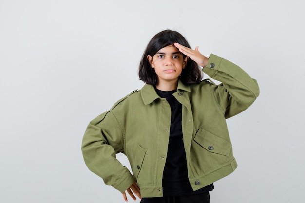 Teen girl showing salute gesture in t-shirt, green jacket and looking confident , front view.